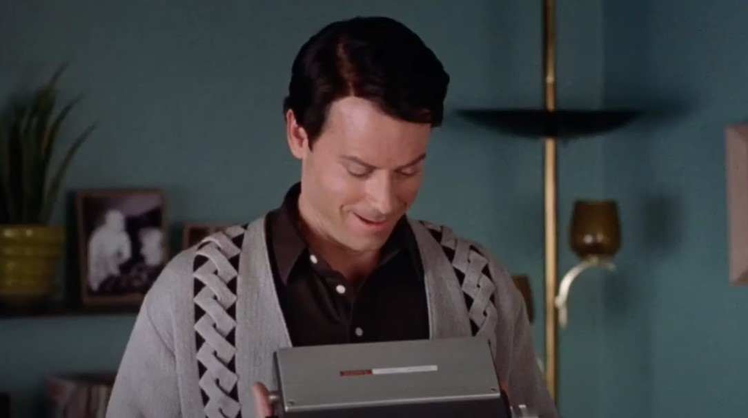 Greg Kinnear  as  Bob Crane looking at a video recorder device in gray sweater