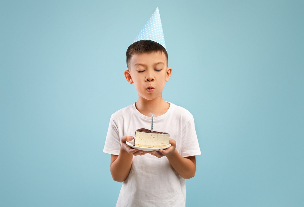 Cute Little Asian Boy In Party Hat Blowing Candle On Birthday Cake in white t-shirt