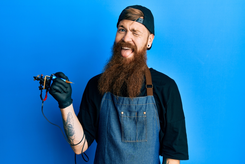Redhead man with long beard tattoo artist wearing professional uniform and gloves