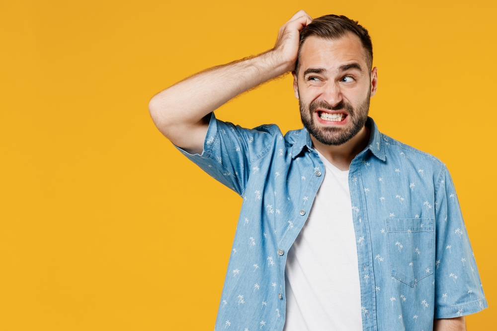 Young puzzled embarrassed Caucasian man wearing blue shirt and white t-shirt scratching his head