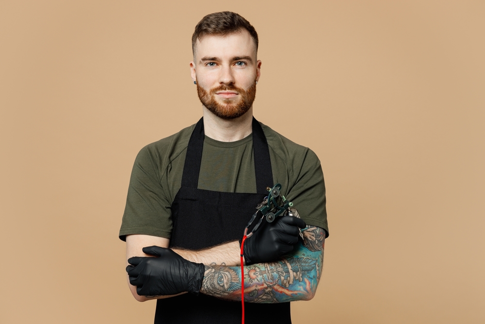 Confident tattoo master artist, tattooed man in green t-shirt and apron hold his hands crossed