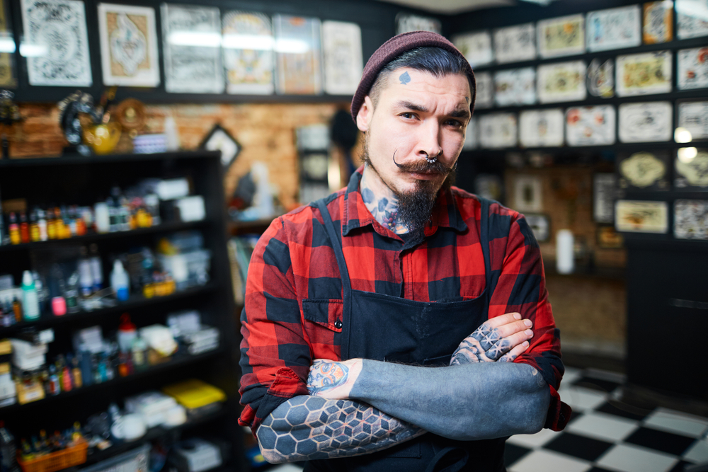 Young bearded hipster tattoo artist in plaid shirt and a hat with tattoos on arms