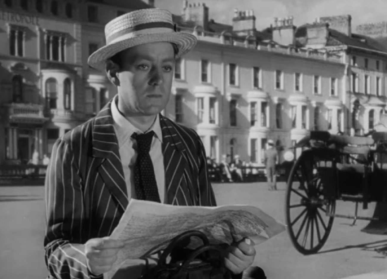 Screenshot of Alec Guinness wearing suit and white hat is reading a paper - from The Promoter (1952)