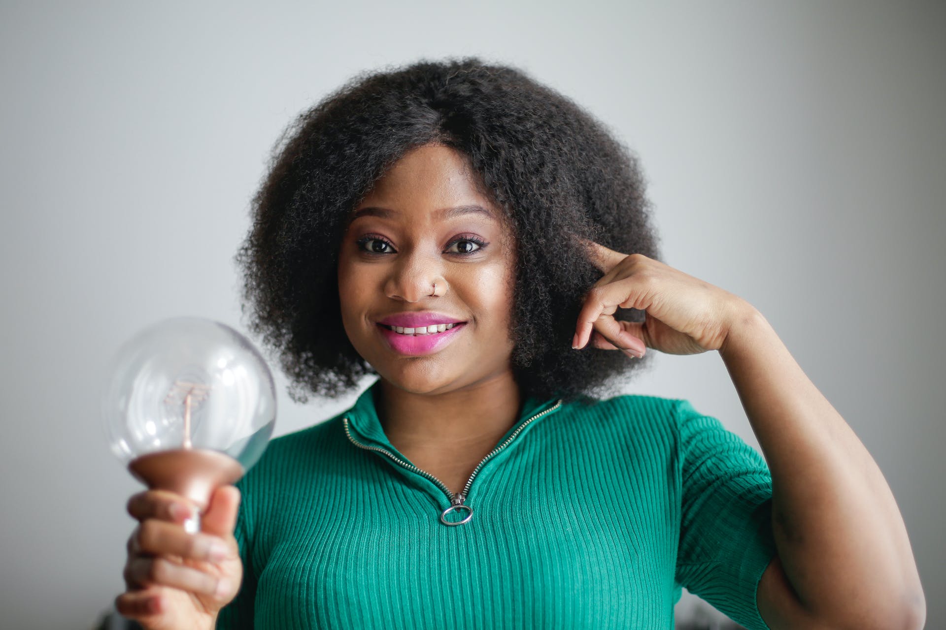 positive young lady wearing green outfit holding light bulb in hand pointing to her head