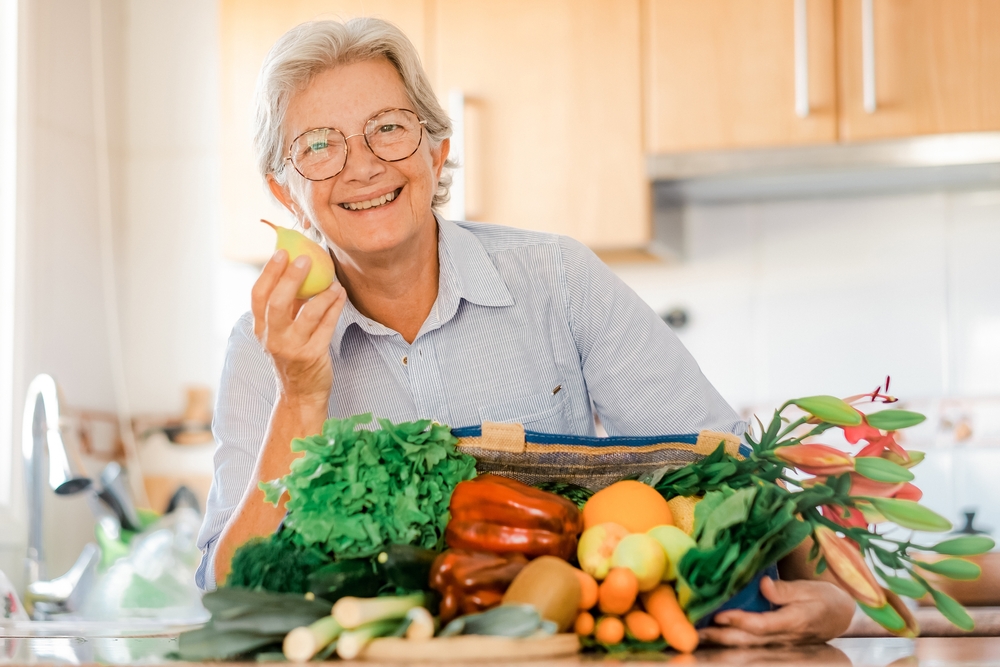 Smiling senior woman in home kitchen with a bag full of vegetables and fruit