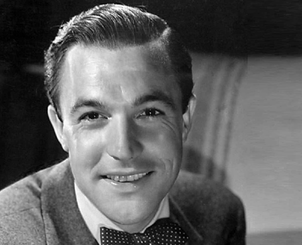 B&W photo of Gene Kelly wearing grey suit with white shirt and tie, smiling to the camera