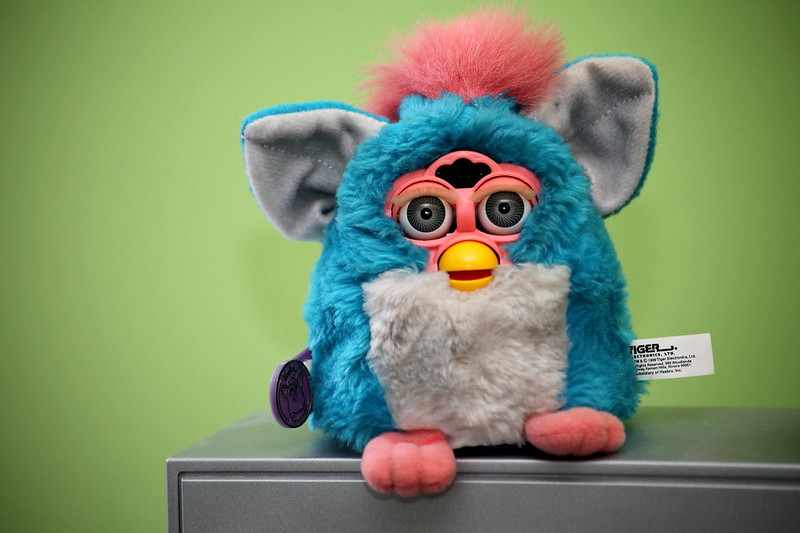A blue and pink Furby sits on a grey surface against a bright green background