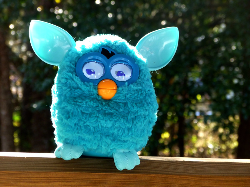 A bright blue Furby sits perched on a wooden railing, with a lush green background of trees
