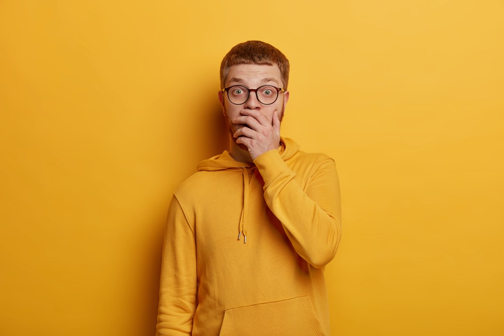 Excited speechless young man covers mouth and gasps from great wonder, hears stunning news, wears transparent glasses and hoodie