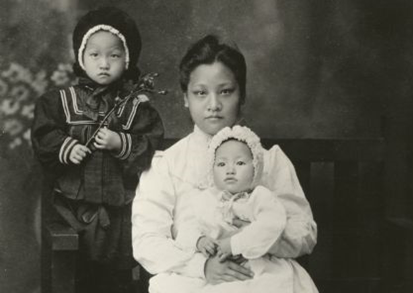 Anna May Wong seated in her mother's lap with her older sister standing, c. 1905.