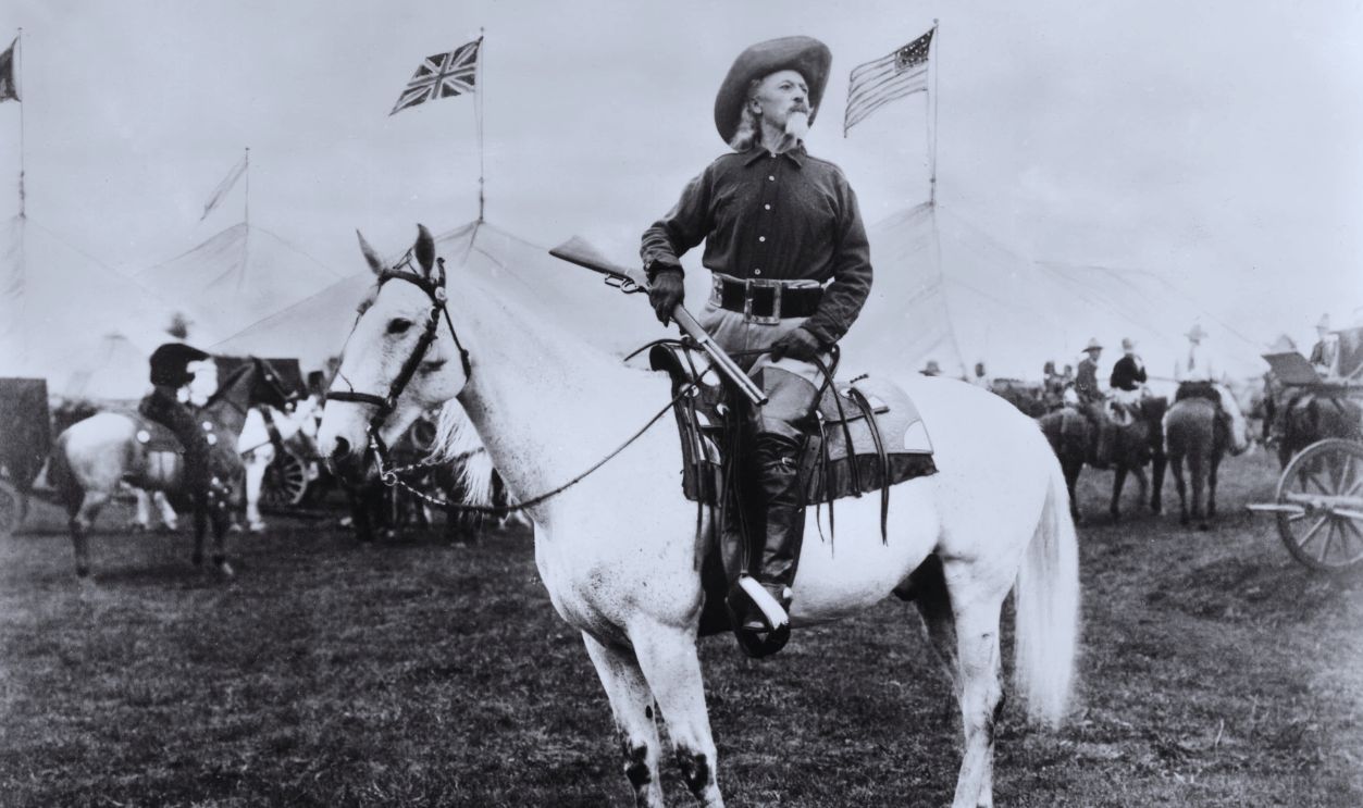 Gettyimages - 3207366, Frontiersman circa 1900: William 'Buffalo Bill' Cody (1846 - 1917) American entertainer, sitting on horseback and holding a rifle, looks off into the distance as British and American flags fly around him. Tents for his Wild West show are in the background.