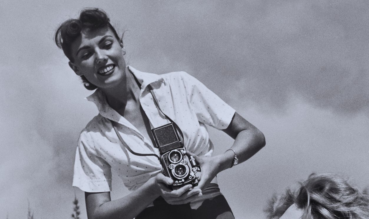 Gettyimages - 2164604800, Bunny Yeager, American Photographer & Pin-Up Model American photographer and pin-up girl Bunny Yeager, wearing a tie-front polka dot shirt and dark shorts, using a Rolleiflex Compur-Rapid medium format camera stands over a female model wearing a bikini, who looks over her shoulder toward Yeager during a shoot on the seafront in Miami, Florida, circa 1955.