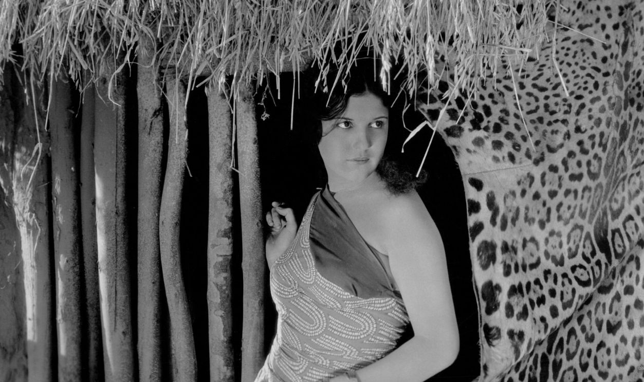 Publicity portrait of silent actor Ann Rork (1908-1988), standing in front of a hut and beside a jaguar fur, as 'Kameela' in the film 'The Notorious Lady', 1927, United States. 