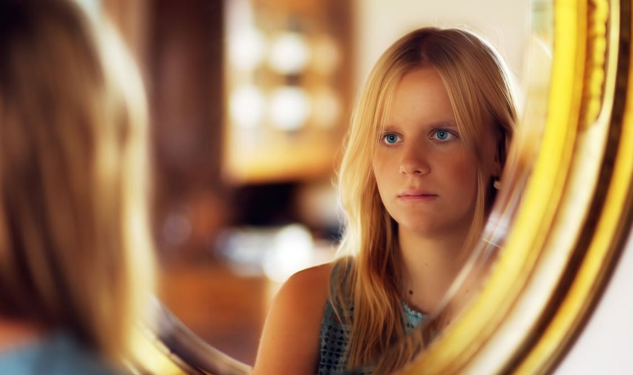Blonde woman gazing seriously at her reflection in the mirror.