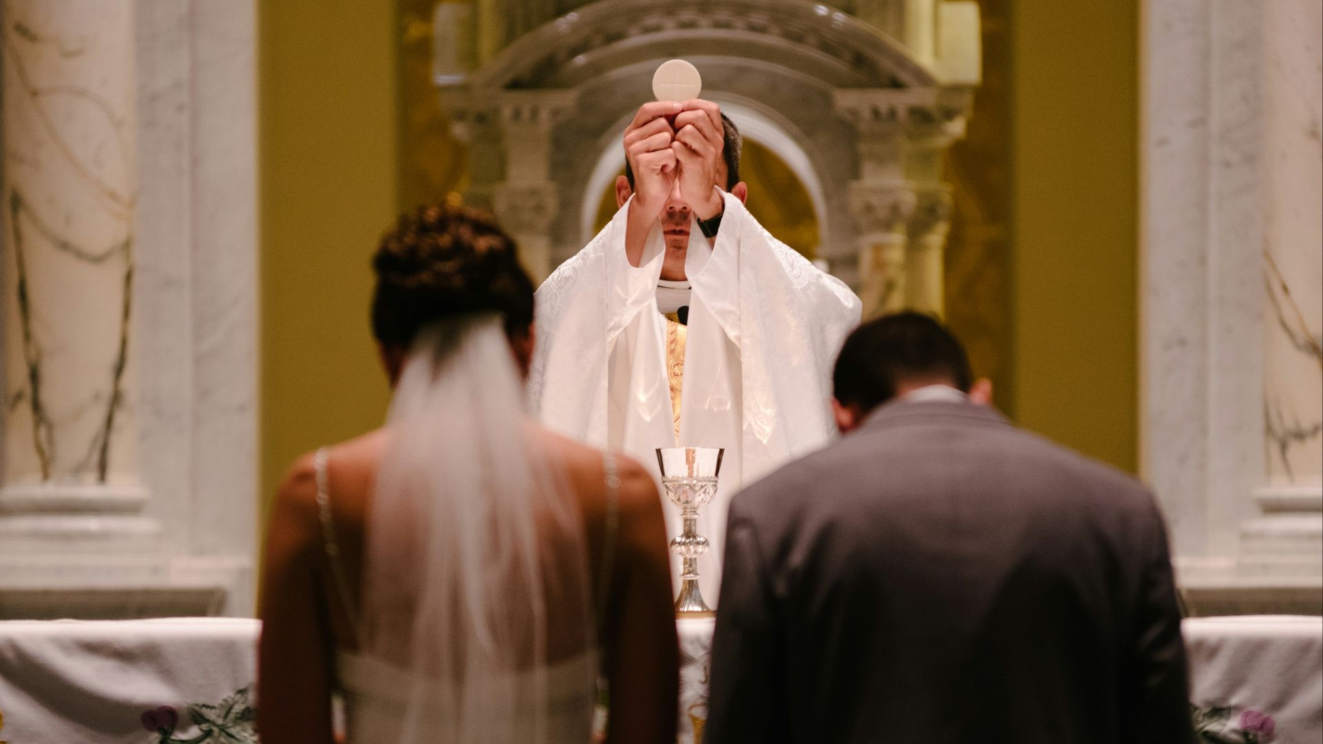 groom and bride kneeling in front of priest raising The Holy Sacrament