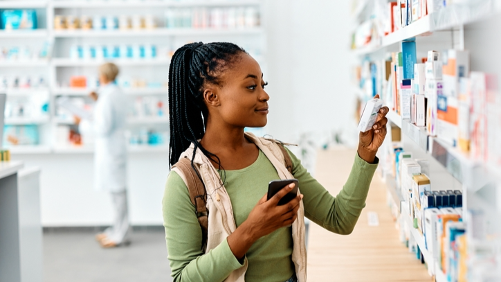 Young African American woman using cell phone while looking for a medicine in pharmacy wearing green top