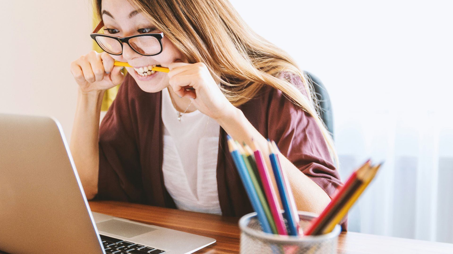 woman biting pencil while sitting on chair in front of computer during daytime