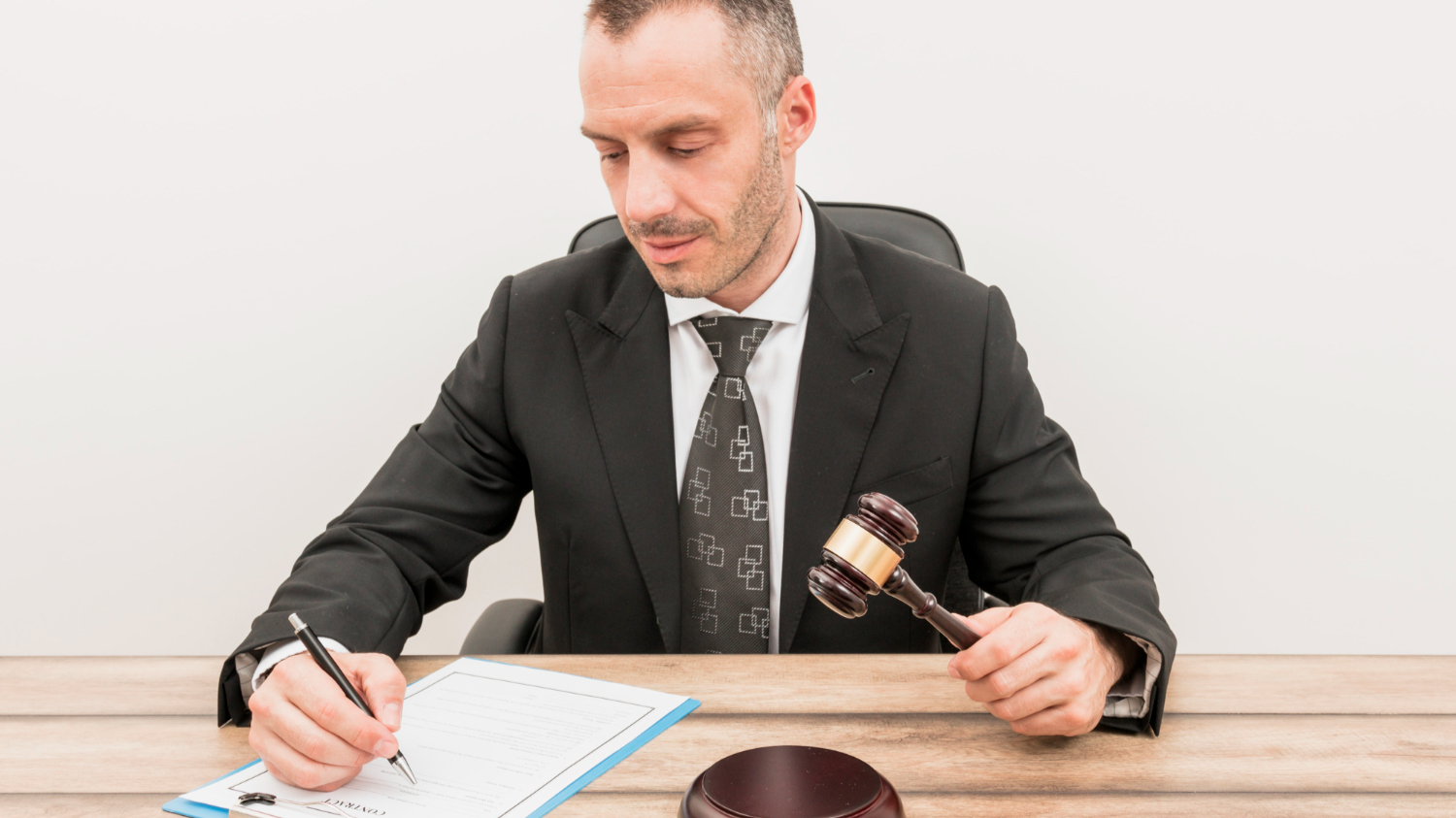 Man is holding a pen ,seating on the bench with a judge hammer in his hand.