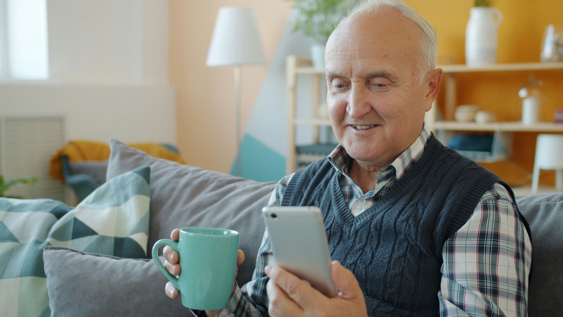 Smiling senior man holding mug and phone on couch.