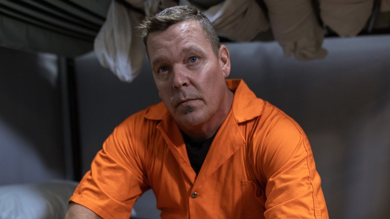 Man is seating in jail cell and looking at camera.