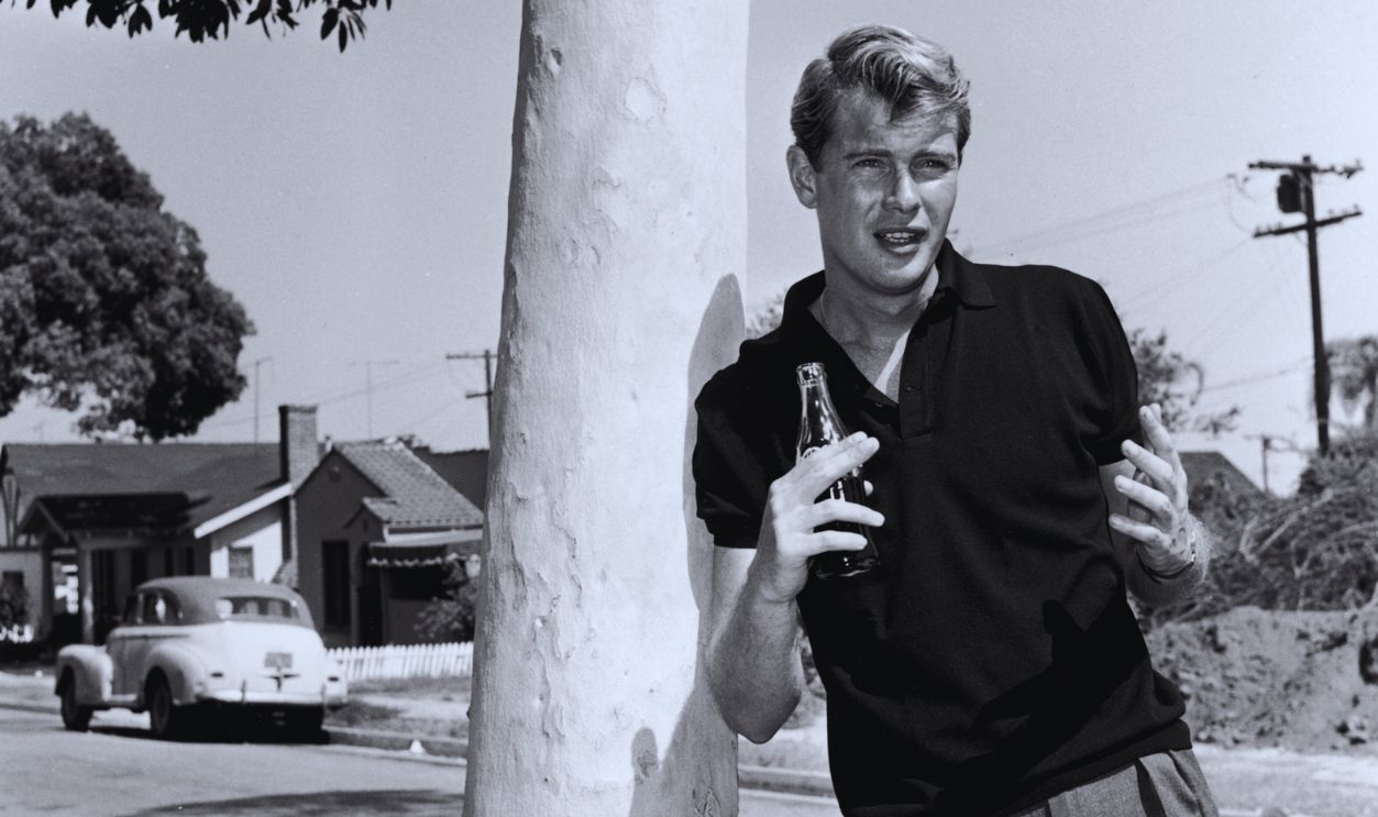 Gettyimages - 3202693, Troy Donahue 1956: Portrait of American actor Troy Donahue (1936 - 2001) leaning against a tree and gesturing with his hands while holding a Coca-Cola bottle on a residential street, California. Donahue wears a dark polo shirt and pleated pants. 