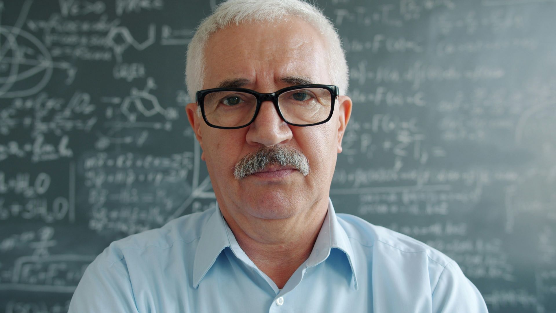 Older man with glasses in front of a chalkboard.