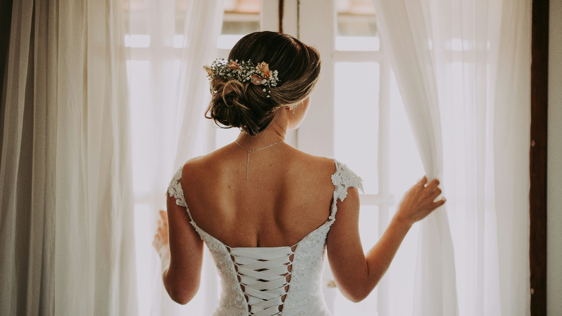 woman in wedding dress standing near window looking outside while holding the curtains
