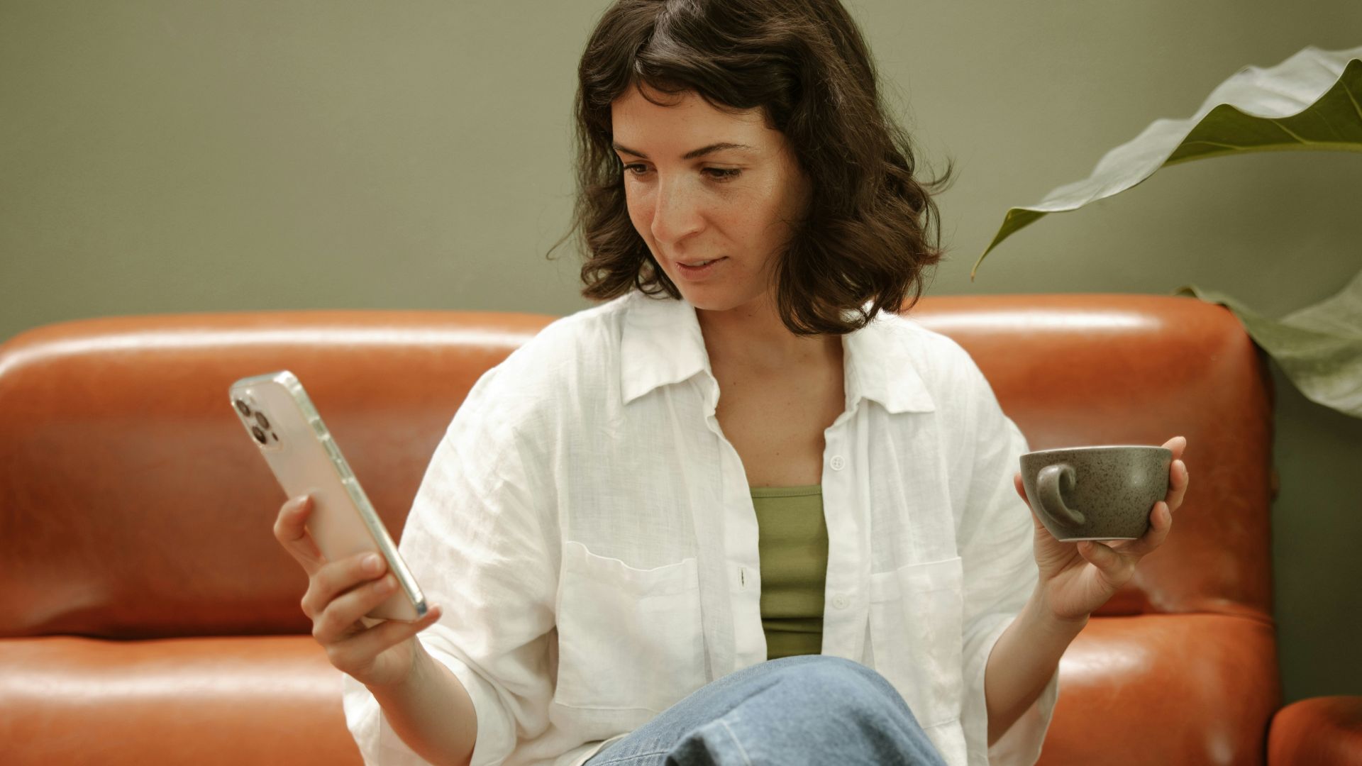 a woman sitting on a couch holding a cup of coffee and a cell phone