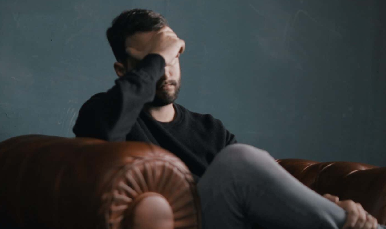 A Man Holds His Head While Sitting On A Sofa