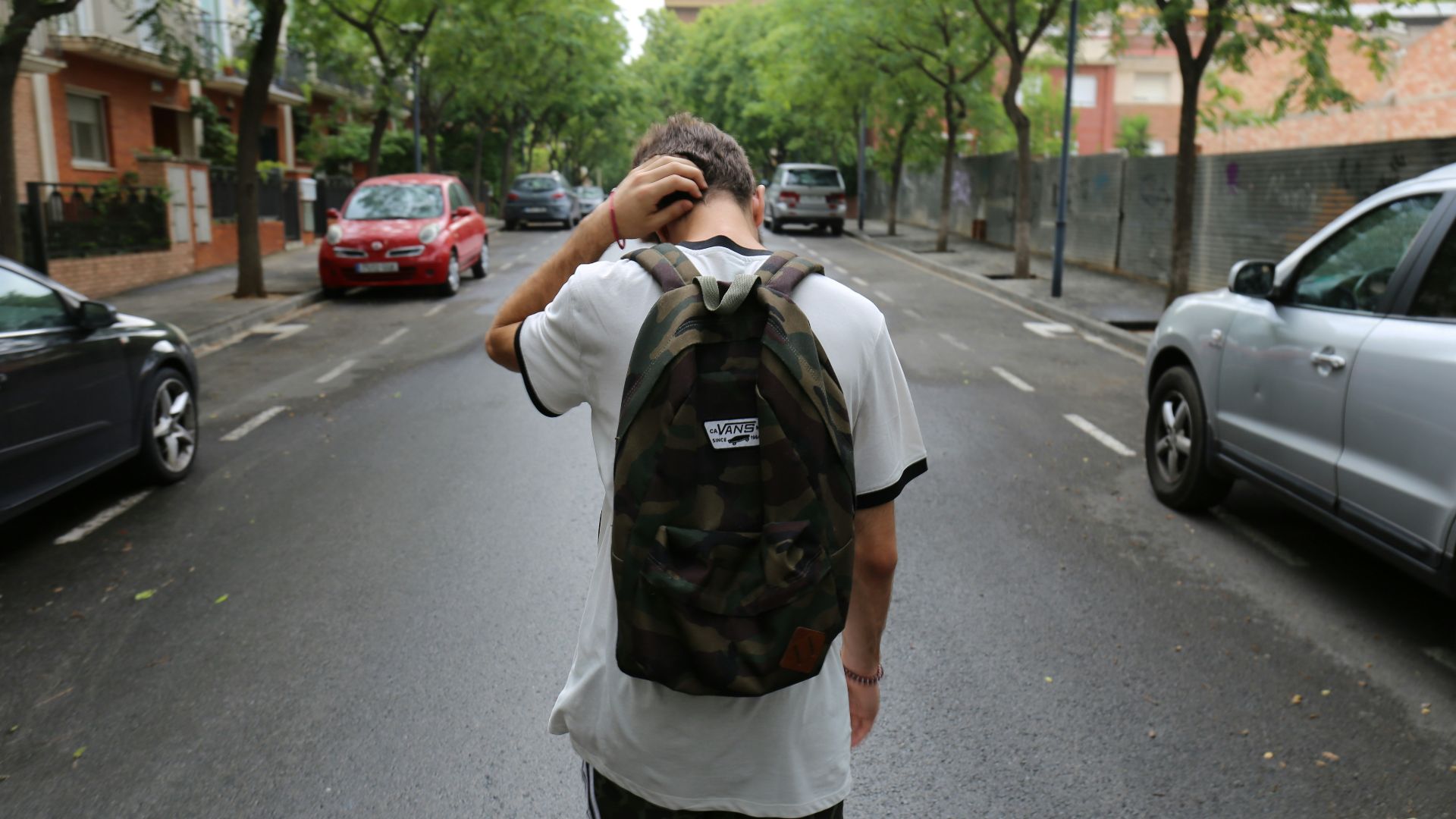 boy wearing white shirt and black shorts carrying backpack standing on black concrete road between vehicles and trees during daytime