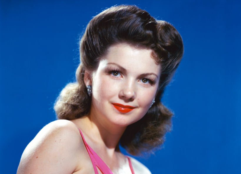 US actress Anne Baxter wearing a pink halterneck top in a studio portrait, against a blue background
