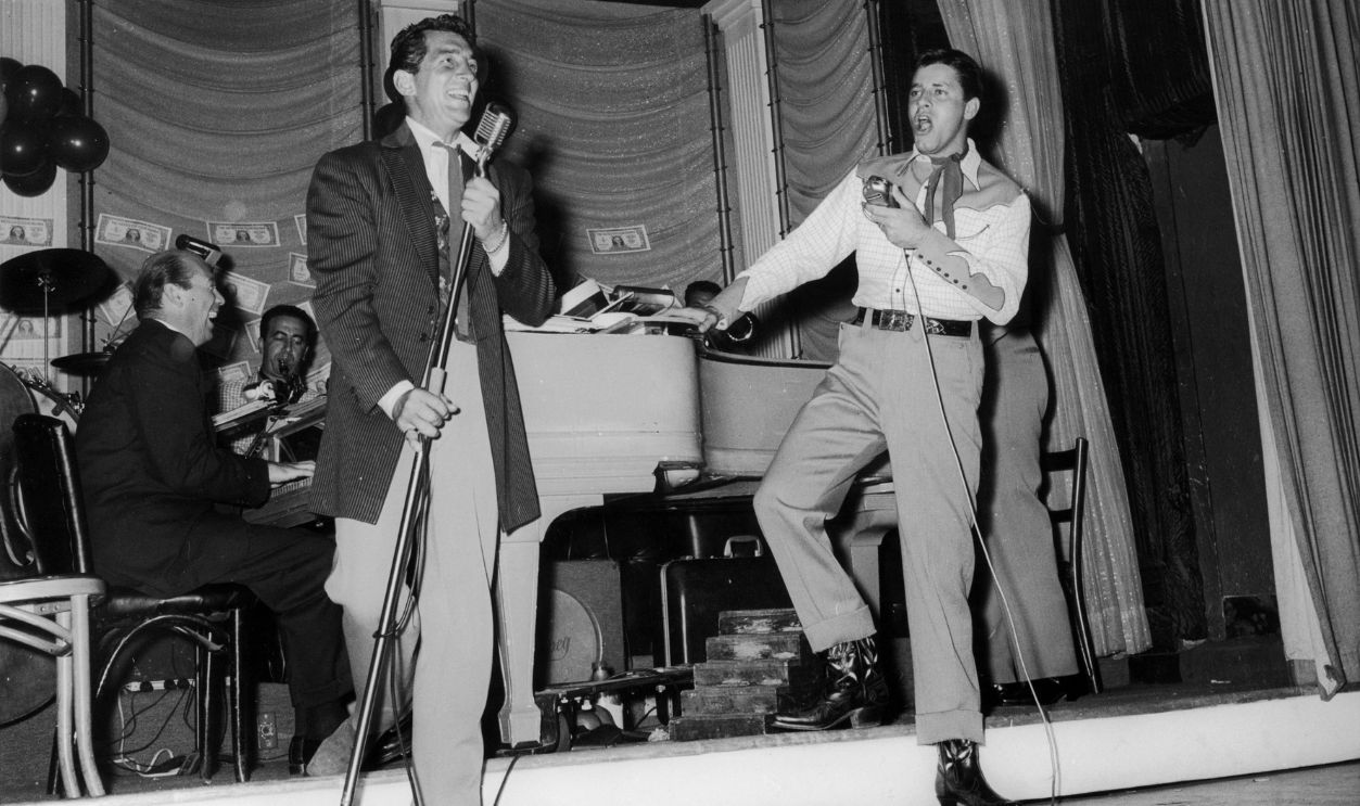 Gettyimages - 3241126, Charity Comics circa 1955: American comic team Jerry Lewis (right) and Dean Martin (1917 - 1995) sing into microphones onstage while performing at a SHARE Boomtown party for charity. Behind them sits a piano player and other musicians.