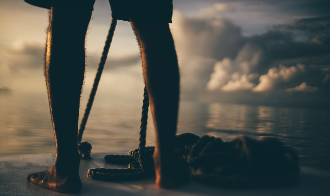 Barefoot on boat at sunset - stock photo Tilt-shift low-key shot of a person standing barefoot on a boat deck, holding a rope. The blurred background features a calm sea and dramatic clouds at sunset, creating a serene atmosphere