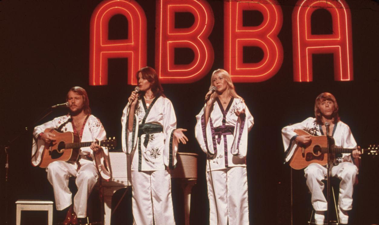 Swedish pop group ABBA (L-R: Benny Andersson, Frida Lyngstad, Agnetha Faltskog, Bjorn Ulvaeus), wearing similar white Asian-influenced costumes, perform for the television program Midnight Special with a neon 'ABBA' sign in the background.