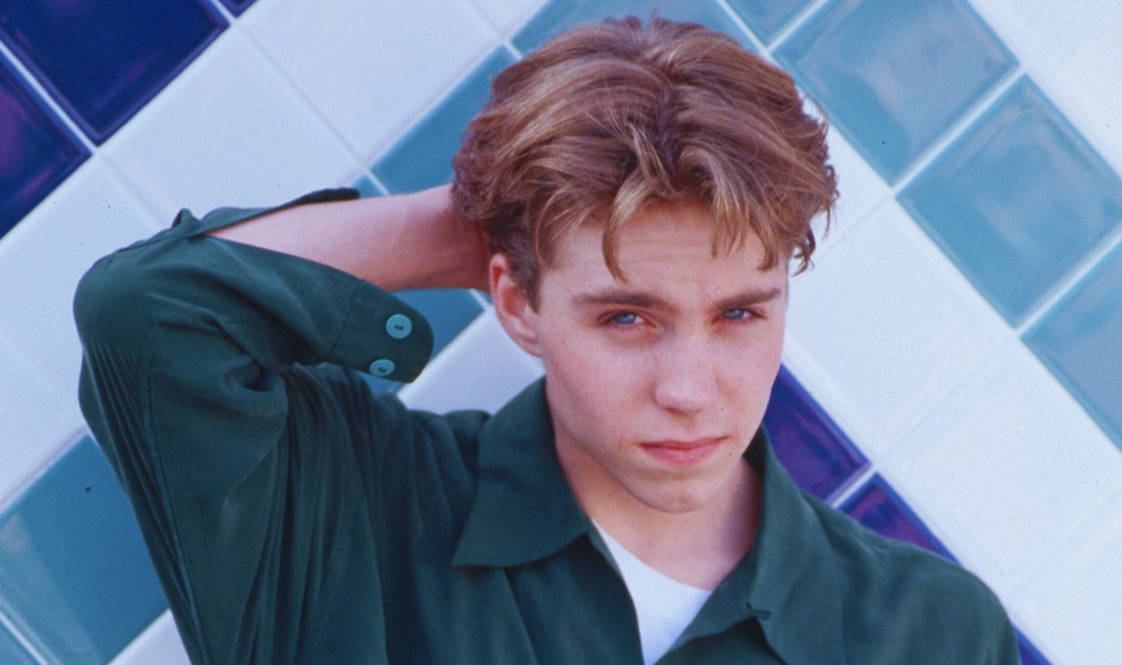 Gettyimages - 635762347, Teen Actor Jonathan Brandis (Original Caption) : 1994-Los Angeles,CA-Jonathan Brandis Posing in front of a white and blue tile wall. He has one hand behind his head and the other is behind his back.