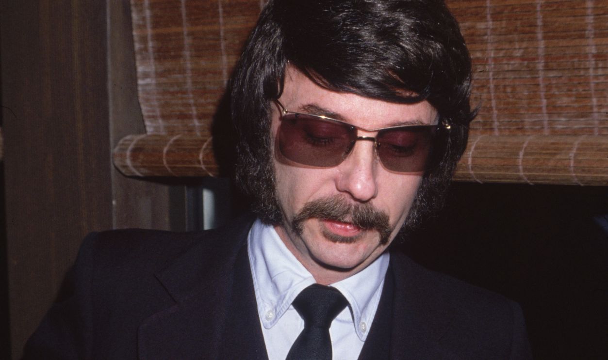 circa 1975: Headshot of American music producer Phil Spector in dark glasses and wearing a badge that reads, 'Back to MONO.'