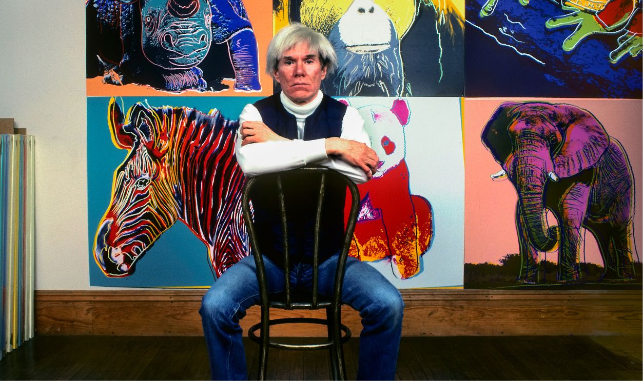 American Pop artist Andy Warhol sits in front of several paintings in his 'Endangered Species' at his studio, the Factory, in Union Square, New York, New York, April 12, 1983.
