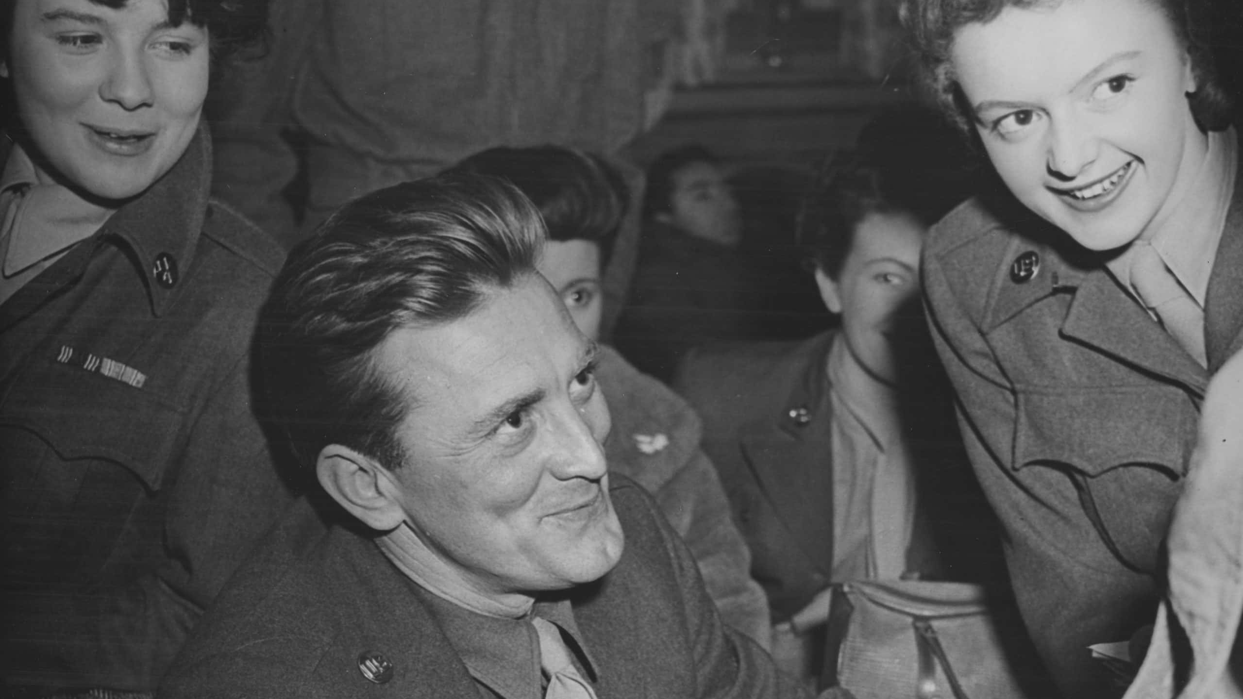 Kirk Douglas, American actor, pictured smiling while smoking a cigarette, circa 1940. Two female admirers stand either side of Douglas are wearing military dress.