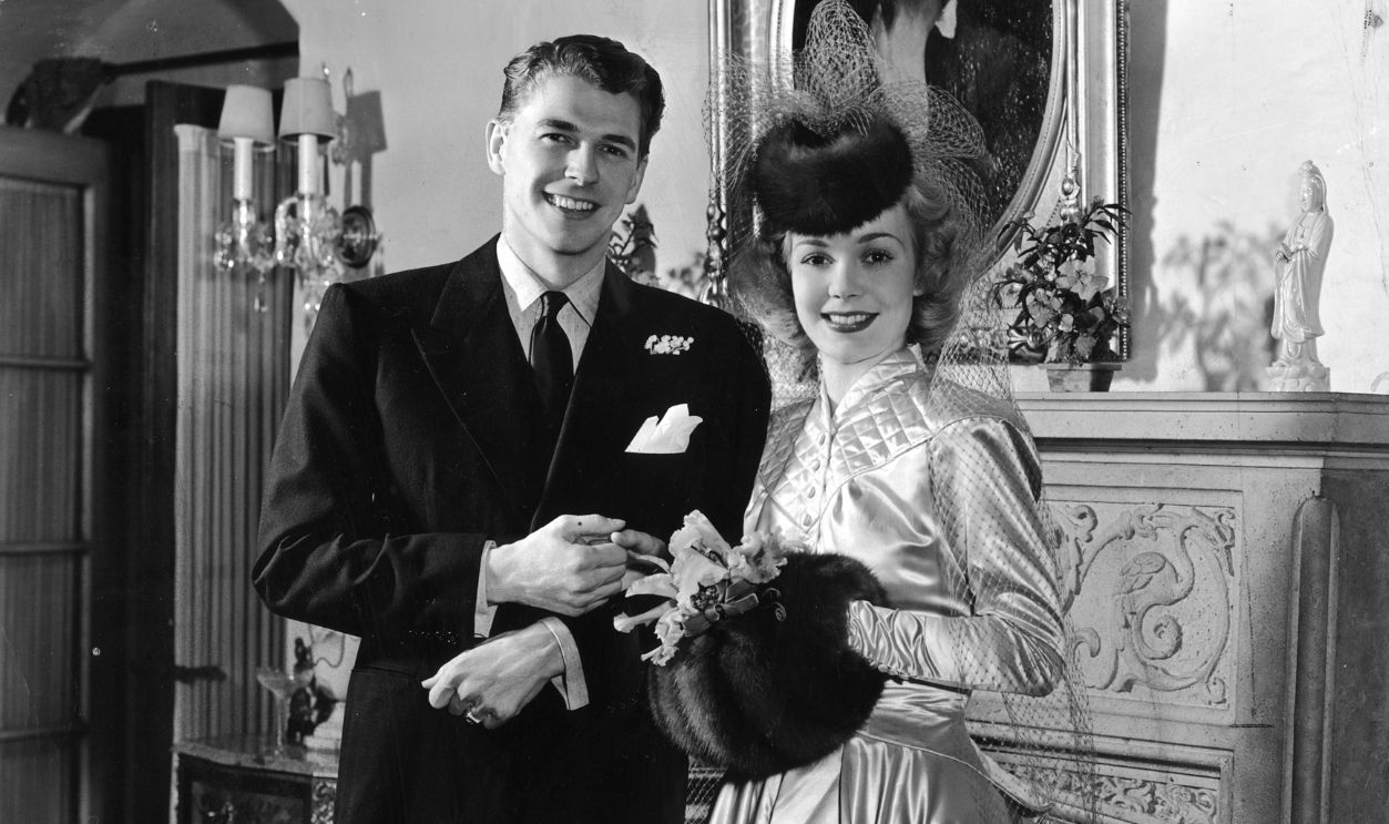 Full-length portrait of American actor Ronald Reagan with his first wife, American actor Jane Wyman holding hands in front of a stone fireplace on their wedding day. Wyman is wearing a satin dress with long sleeves and a high collar. Her hands are in a muff. Reagan is dressed in a black suit with a white handkerchief in the breast pocket and a small pin of flowers under the lapel