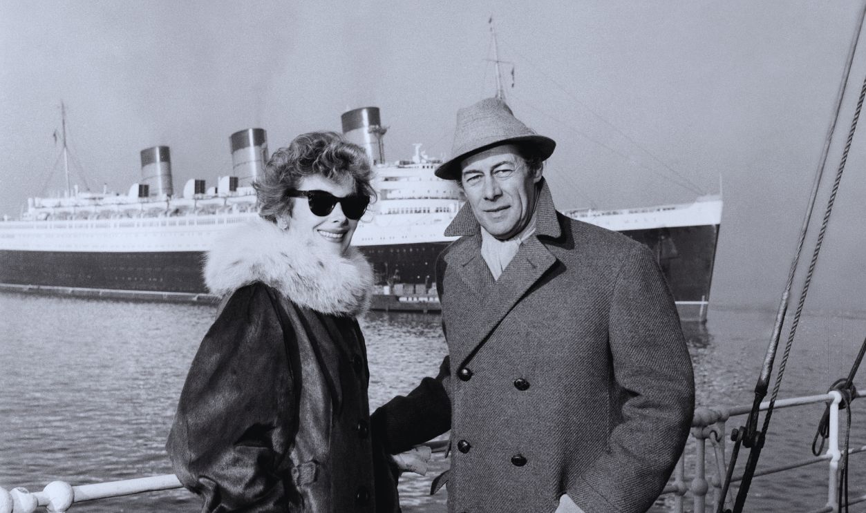 Gettyimages - 2201920866, Kay Kendall, Rex Harrison, 1957 Actress Kay Kendall smiles while waiting to board ocean liner 'RMS Queen Mary' (in background) with her husband, actor Rex Harrison, Cherbourg, France, December 10th 1957.