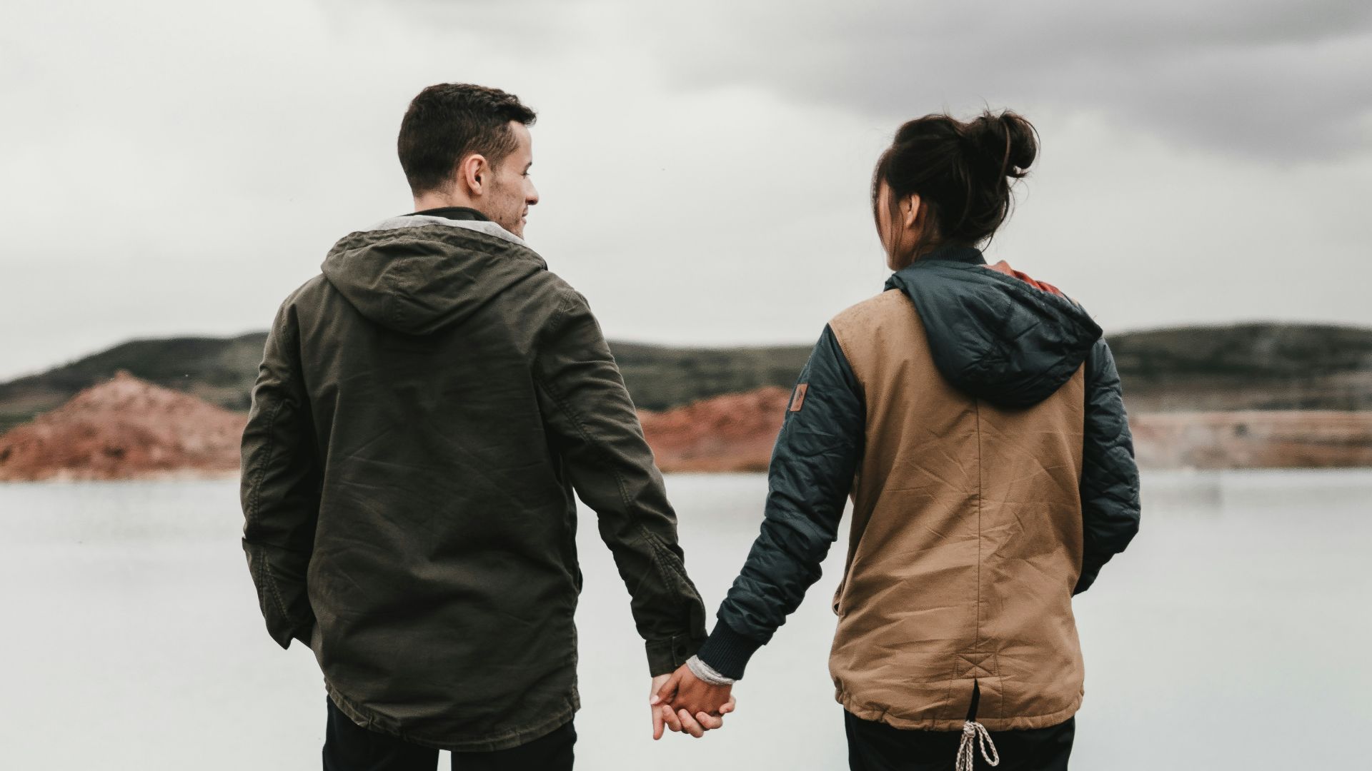 couple holding hand front of body of calm water with mountain distance