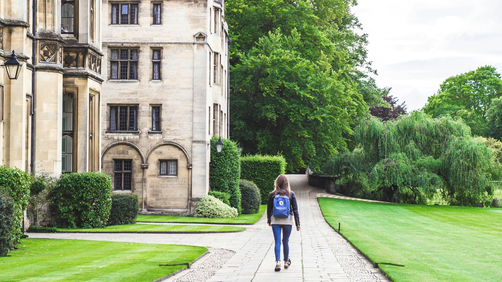 person walking on concrete road beside brown concrete house near green high trees under white sky during daytime