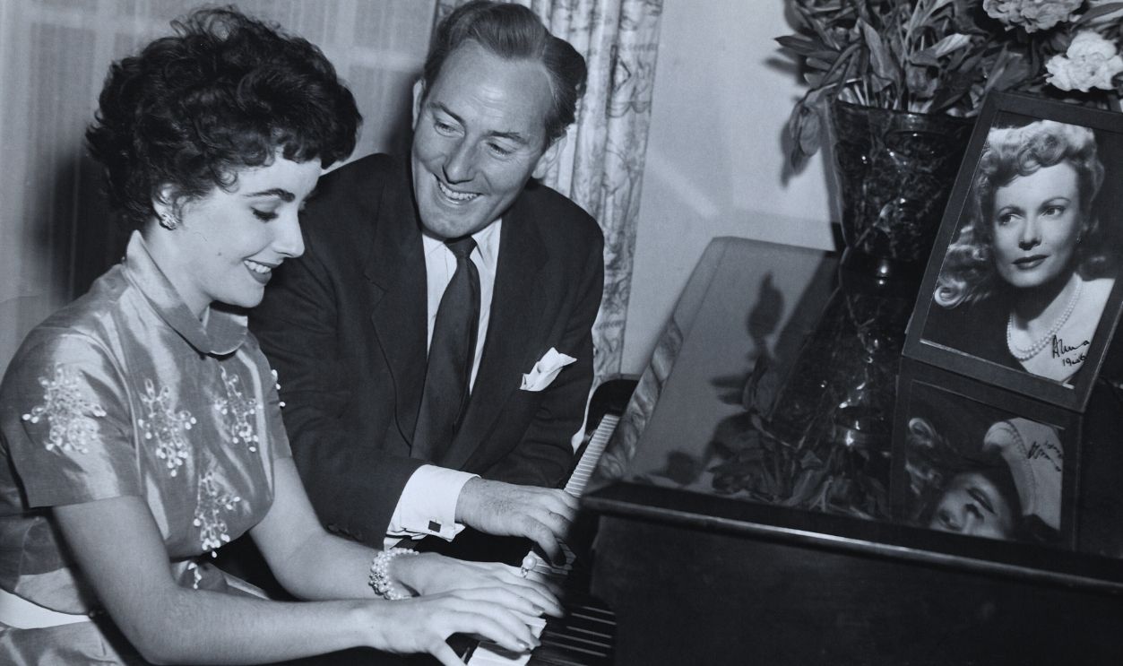 Gettyimages - 514897590, Michael Wilding & Liz Taylor Play Piano (Original Caption) TAYLOR-TICKLING THE IVORIES, Elizabeth Taylor and Michael Wilding show off their pianistic prowess. Pictures of friends are everywhere in the Wilding apartment. The camera portrait atop the piano is that of Anna Neagle, matron of honor at their wedding. Ca. 1950s.
