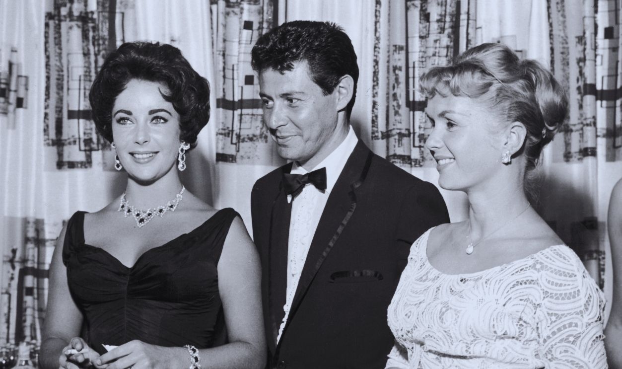 Gettyimages - 3239825, With Eddie Fisher 1958: American singer Eddie Fisher, wearing a tuxedo, stands with arm around his wife, American actor Debbie Reynolds (R) and smiles while looking at British-born actor Elizabeth Taylor, smoking a cigarette, Las Vegas, Nevada. The next year Fisher left Reynolds and married Taylor.