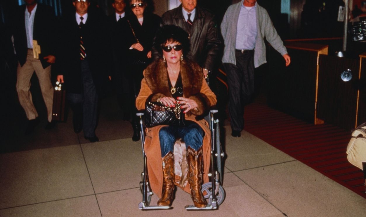 Gettyimages - 2207810818, Elizabeth Taylor A man pushes British born actress Elizabeth Taylor in a wheelchair, circa 1988.