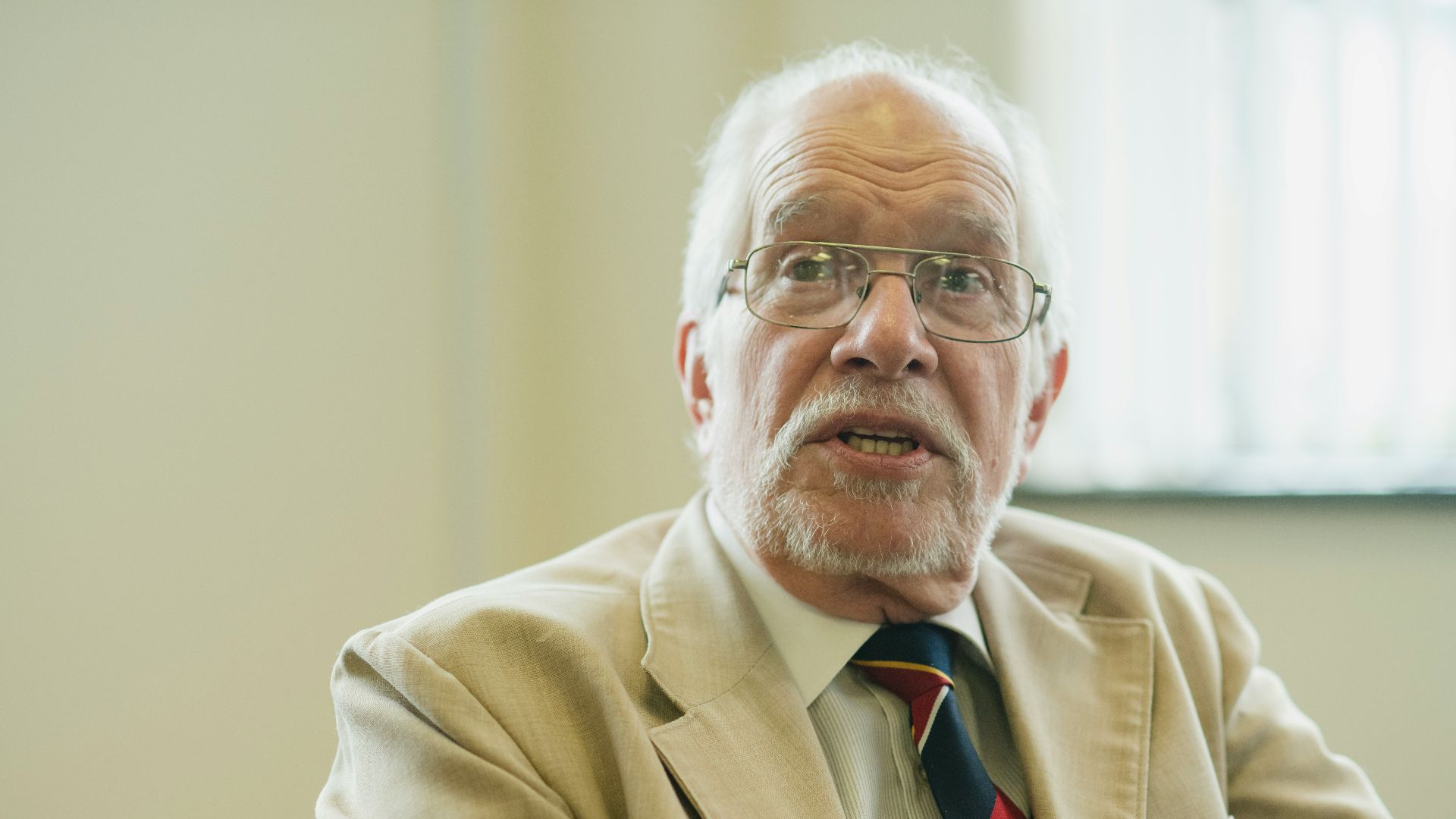 An elderly man in a suit jacket looks at the camera.