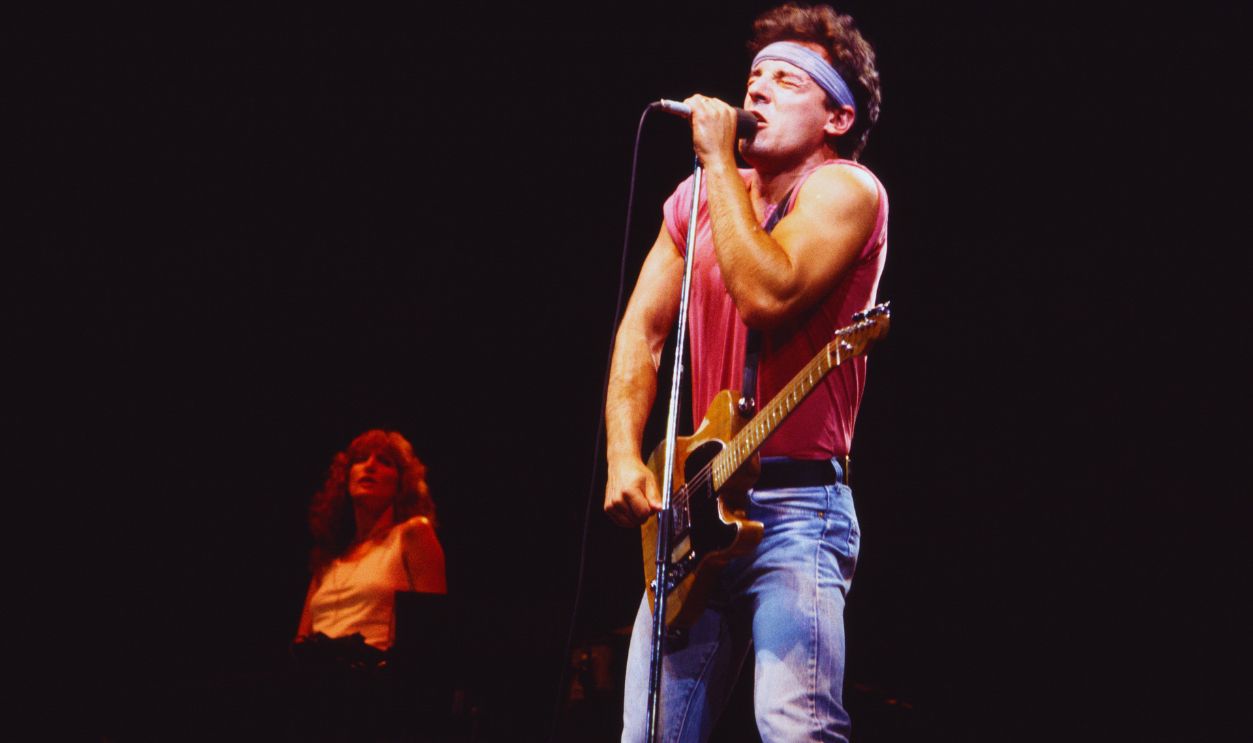 Gettyimages - 1290768016, Bruce Springsteen & The E Street Band Perform At Giants Stadium American Rock musician Bruce Springsteen plays guitar as he performs onstage, with the E Street Band, during the 'Born in the USA' tour, at Giants Stadium, East Rutherford, New Jersey, August 22, 1985. Visible in the background is backing vocalist Patti Scialfa.