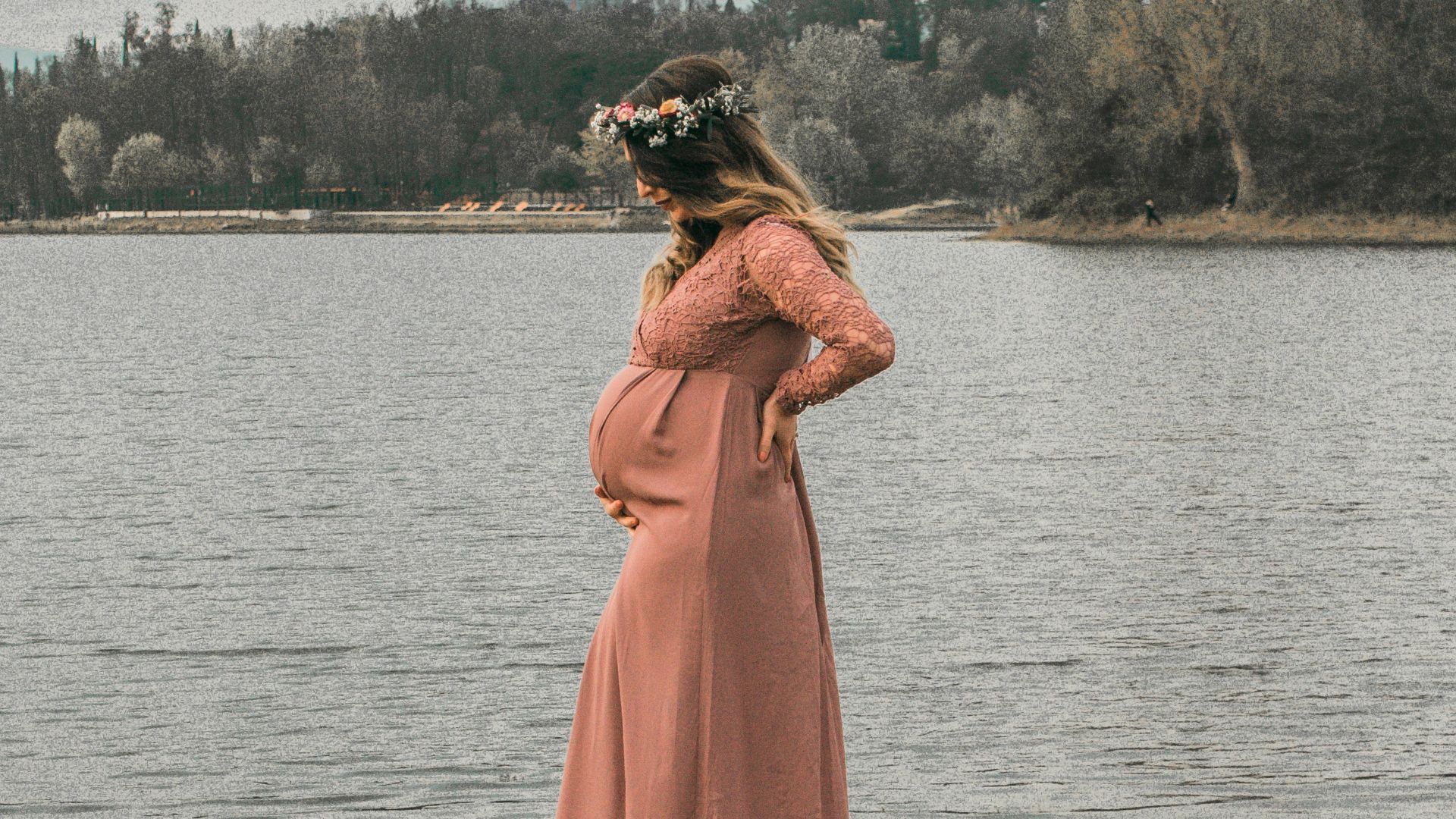 a pregnant woman in a pink dress standing on a rock by a lake