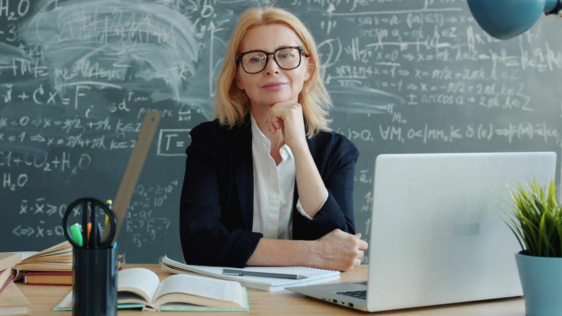 Woman in glasses at desk with laptop and chalkboard.