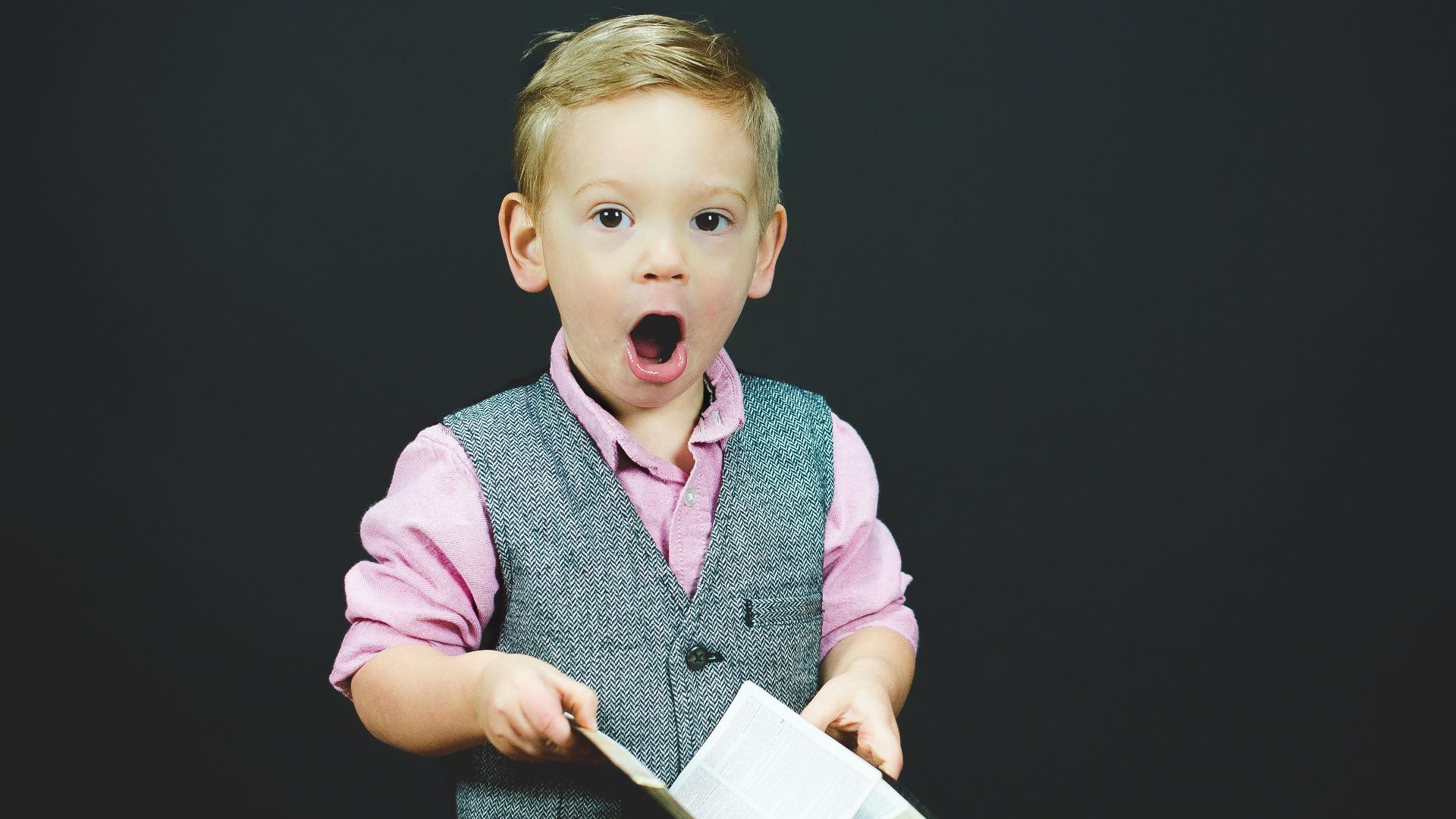 boy wearing gray vest and pink dress shirt holding book